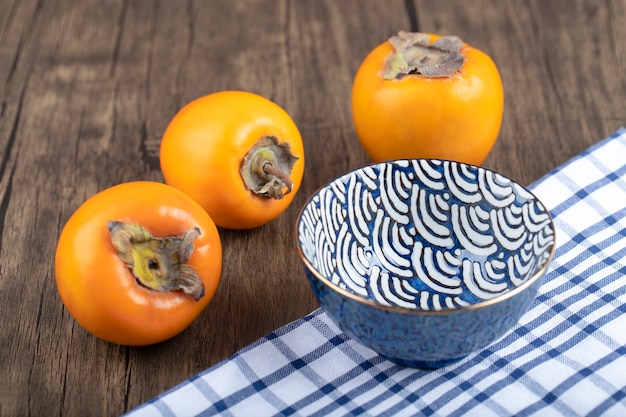 Three persimmon fruits and blue bowl on wooden background. 