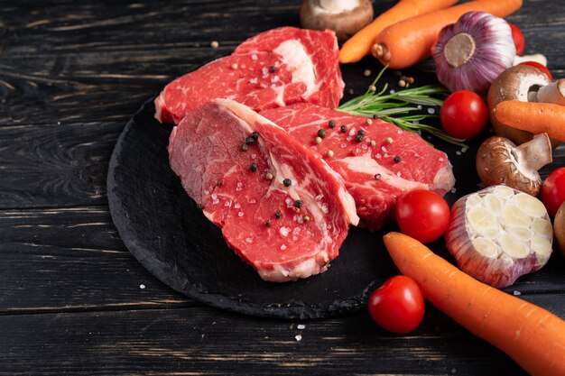 Three pieces of juicy raw beef with vegetables on a stone cutting board on a black wooden table background.