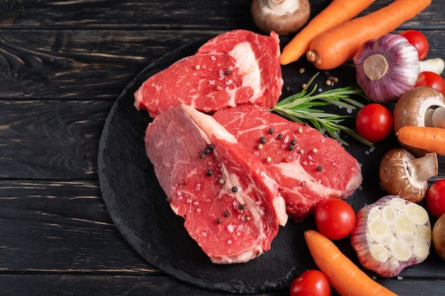 Three pieces of juicy raw beef with vegetables on a stone cutting board on a black wooden table background.