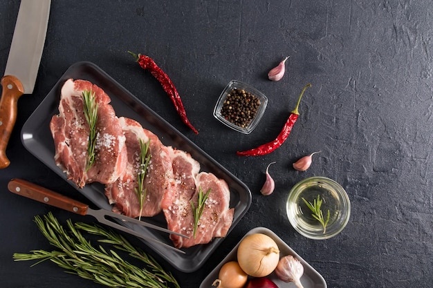 Three pieces of pork neck with rosemary pepper onion garlic on a dark tray a knife and a fork on a black background for cooking top view