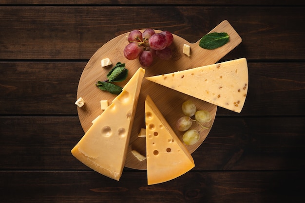 Three pieces of yellow swiss cheese with holes and a branch of red grapes on a cutting board against a wooden surface, top view