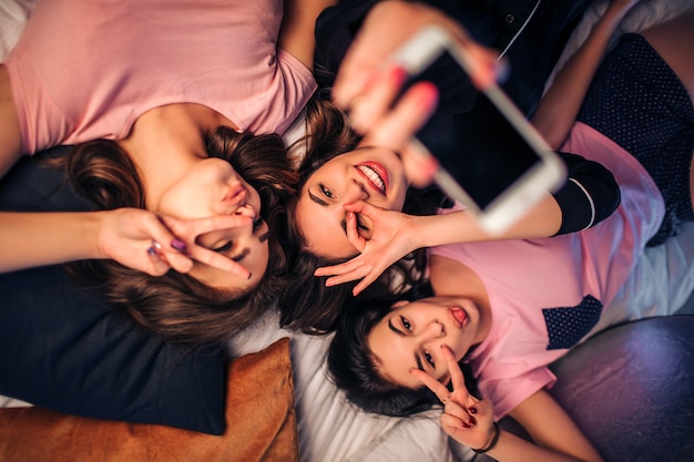 Three playful young women lying on bed. They pose and make different poses. women in middle hold white phone in hand.