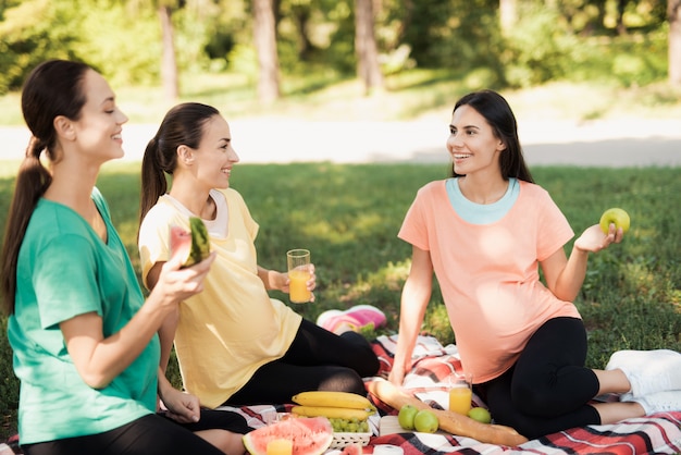 3人の妊婦が公園でのピクニックのために敷物の上に座っています。