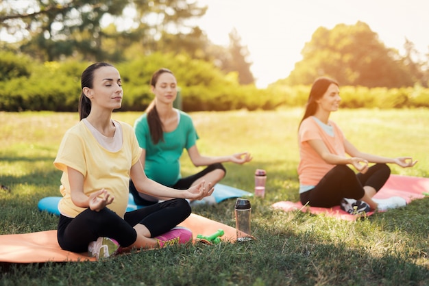 Three pregnant women sit on yoga mats in a lotus pose 