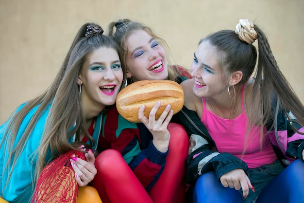 Three pretty girls dressed in the style of the nineties are sitting on the steps and sharing a roll.