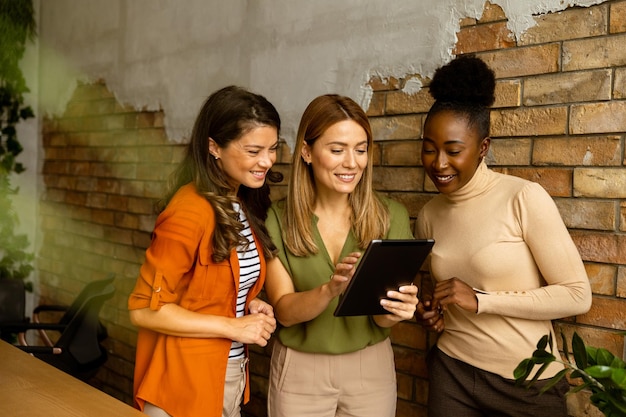 Three pretty young business women with digital tablet standing by the brick wall in the industrial style office