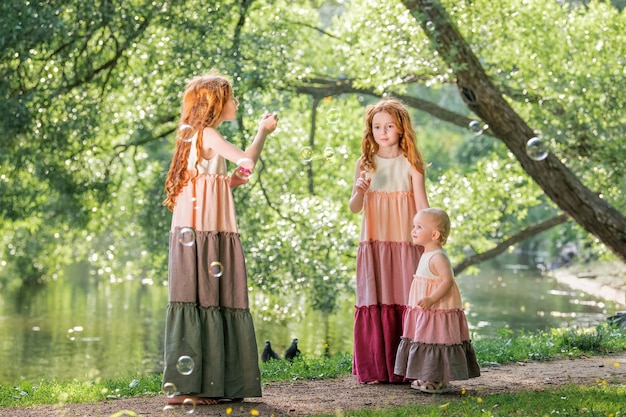 Three red-haired sisters in long linen dresses blow bubbles in the park on a sunny summer day.