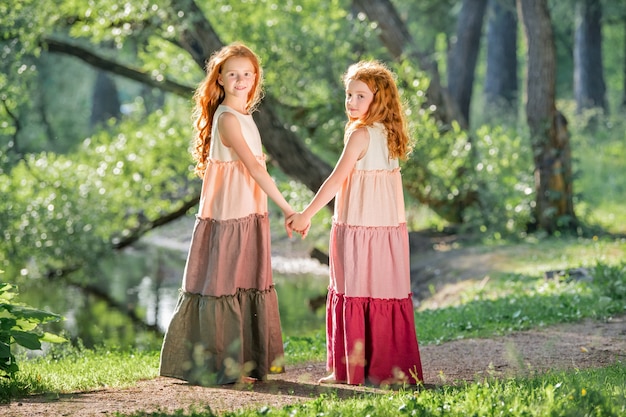 Three red-haired sisters in long linen dresses blow bubbles in the park on a sunny summer day.