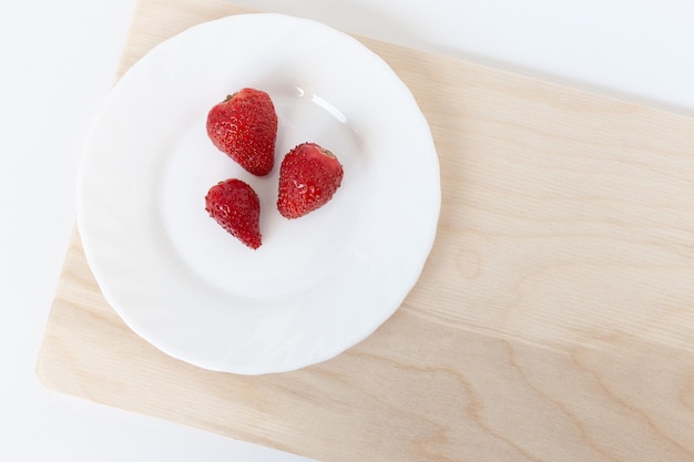 Three red strawberries on a white plate on the brown wooden cutting board.