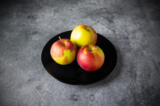 Три красно-желтых спелых яблока на черной тарелке на сером столе