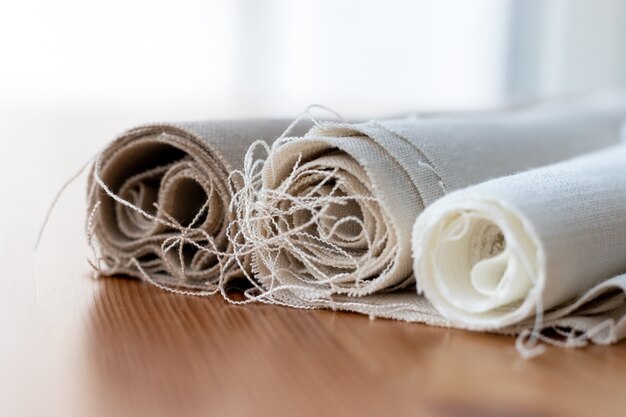 Three rolls of natural linen fabric in different colors with protruding threads on a wooden table.