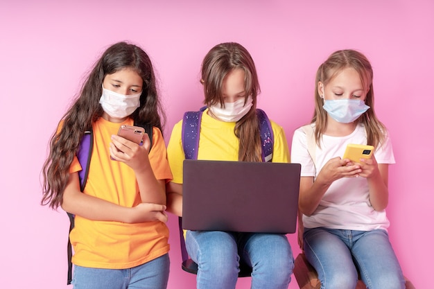 Three schoolgirls in medical masks are holding smartphones and a laptop