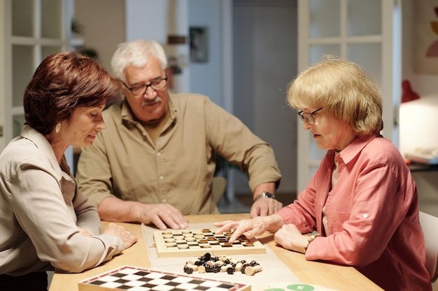 Three senior friends gathered by table in living room for game of chess