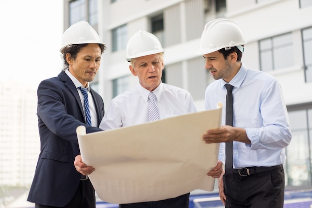 three serious diverse business people wearing helmets, holding blueprint