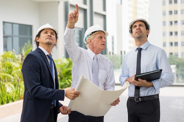 three serious diverse business people wearing helmets, holding blueprint