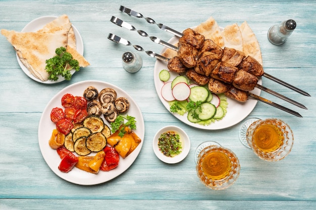 Три порции аппетитного домашнего шашлыка, нарезанные огурцы и редис, овощи, барбекю, бокалы вина, вид сверху на синем деревянном фоне