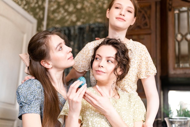 Three sisters beautiful young women ready to celebrate the spring holiday of happy Easter at home decorating dining table with Easter eggs holding in hand vintage countryside fashion style of dress