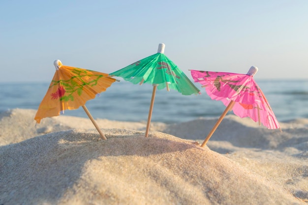 Three small paper cocktail umbrellas stand in sand on sandy beach closeup Small paper umbrellas on sandy shore near sea and sea waves on sunny summer day Leisure Vacation Travel Tourism Concept