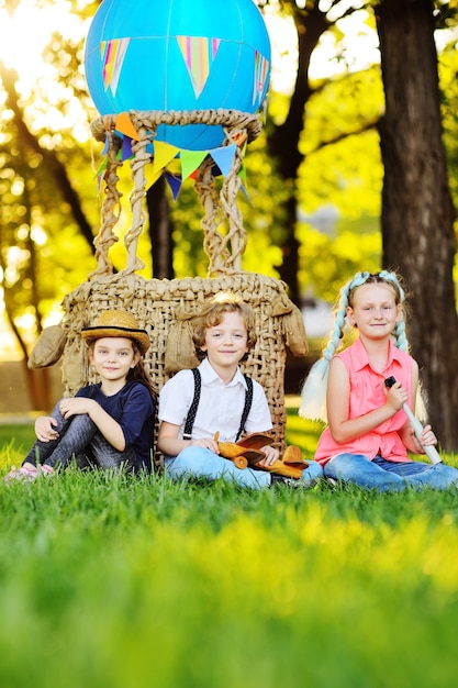 Three small preschool children sit on the grass against the background of a basket of blue balloons and sunlight. Childhood, adventure, vacation.