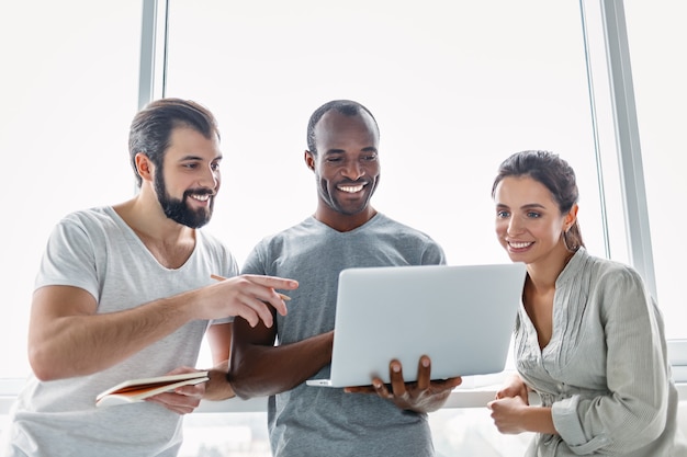 Three smiling business colleagues standing in modern office interior looking at the screen