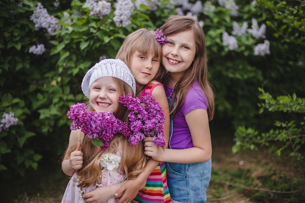 Three smiling European girls stand hugging in a summer garden with lilac flowers in their hands