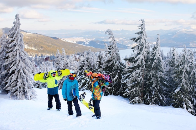 Three snowboarders walking at ski resort Friends climbing to mountain top carrying their snowboards through forest for backcountry freeride and wearing reflective goggles colorful fashion clothes