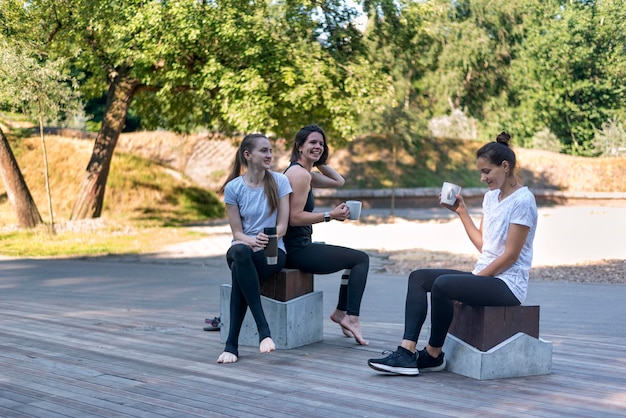 3명의 스포츠 소녀들이 공원에서 차 한잔과 함께 휴식을 취합니다. 여성 그룹은 피트니스 수업 후 휴식을 취합니다