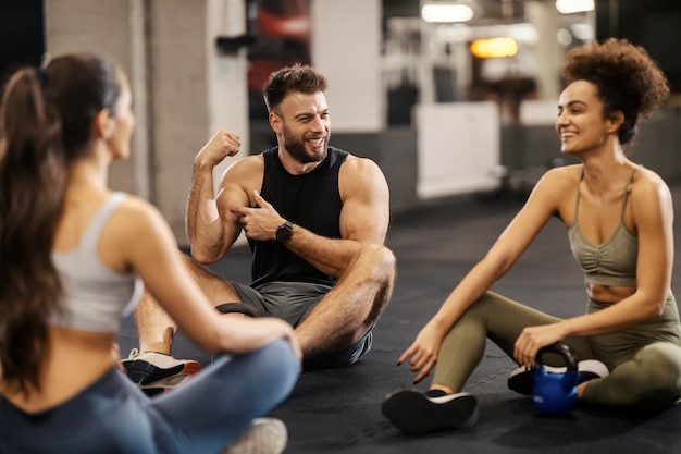 Photo three sporty friends sitting on a gym floor and showing muscles during a break