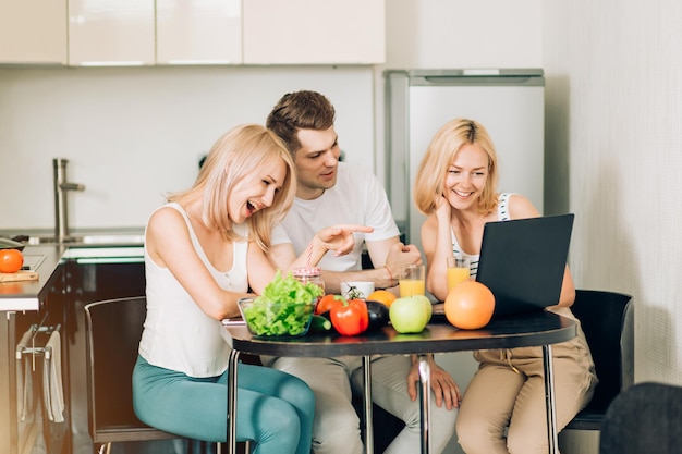 Three students in domestic clothes sitting at the table in kitchen, going to prepare breakfast, watching video recipe on laptop, drinking orange juice. Technology, people concept.