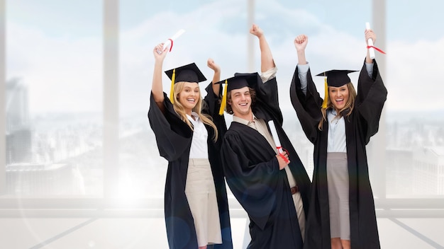 three students in graduate robe raising their arms against bright white room with windows