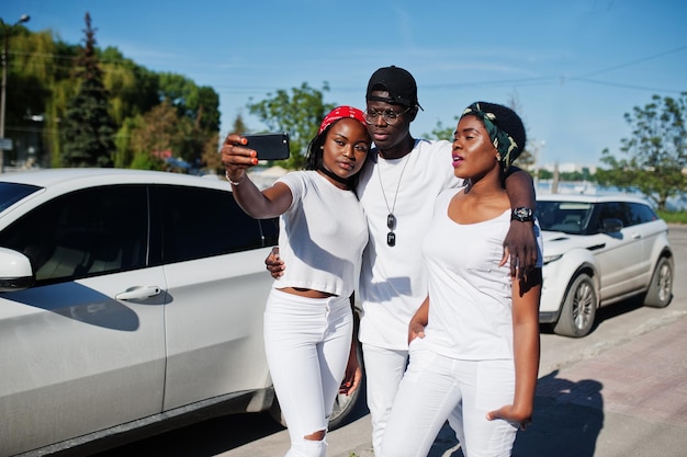 Three stylish african american friends making selfie on phone wear on white clothes against two luxury cars Street fashion of young black people Black man with two african girls