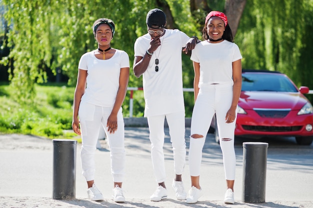 Three stylish african american friends, wear on white clothes against luxury red sport car. Street fashion of young black people. Black man with two african girls.