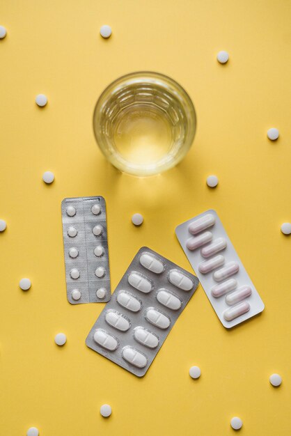 Three tablet blister packs and a water glass on a yellow background They are surrounded by pills