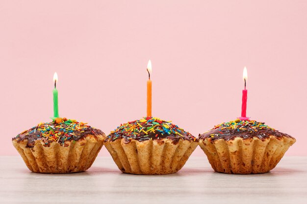 Three tasty birthday cupcakes with chocolate glaze and caramel, decorated with burning festive candles on pink background. Happy birthday minimal concept.