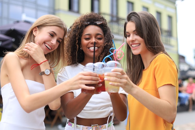 Tre ragazze cool hipster alla moda, amici bevono cocktail sullo sfondo urbano della città.