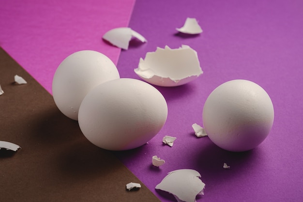 Three white eggs with eggshell pieces on purple, pink and brown colors plain minimal background, angle view, happy Easter day