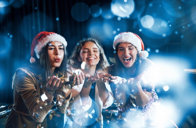 Three women blowing confetti from their hands indoors at holiday time.