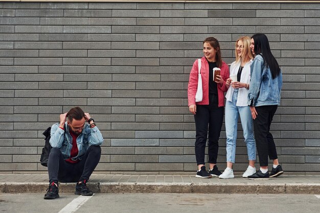 Three women standing and laughing at guy that sitting outdoors near building at daytime