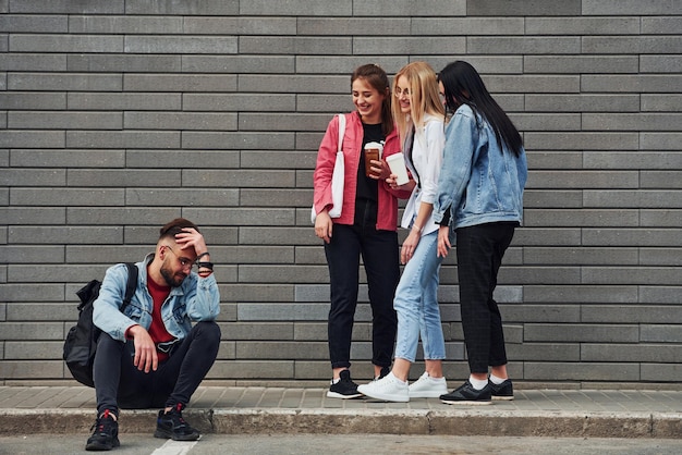 Three women standing and laughing at guy that sitting outdoors near building at daytime