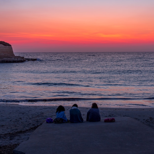 Three women waiting for the sunrise in front of the sea (Puglia region, South of Italy). Concept of frienship, travel and adventure.