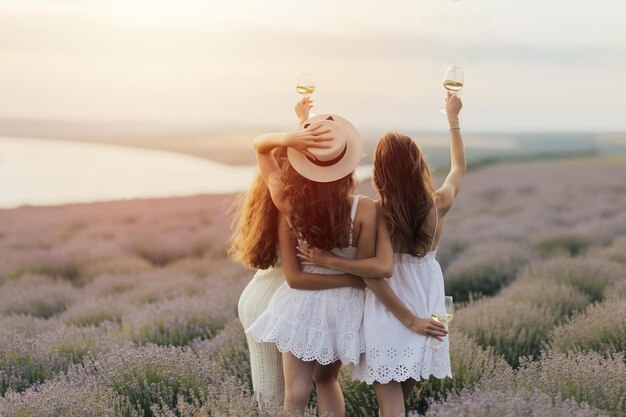 Three women in white dresses are holding glasses of wine in a lavender field