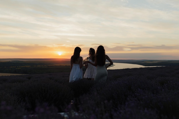 Three women in white dresses stand in a lavender field at sunset.