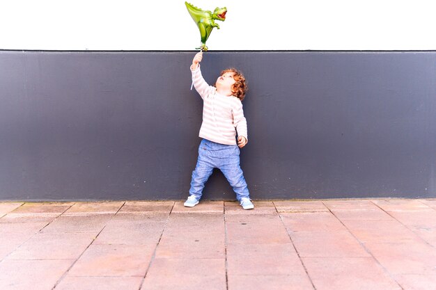 Three year old boy leaning against a white and gray wall playing with a balloon at sunset in spring or summer