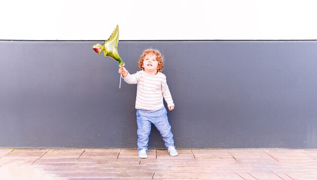 Three year old boy leaning against a white and gray wall playing with a balloon at sunset in spring or summer