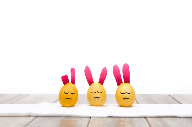 Photo three yellow eggs with funny faces and bunny ears  on a white towel