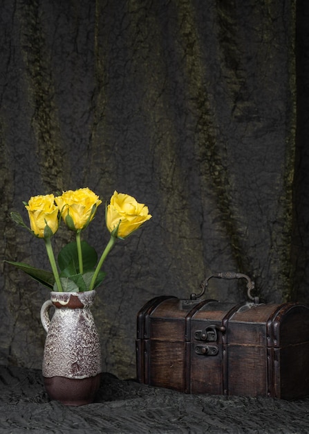 Three yellow roses in a vase on a dark fabric background next to an old wooden chest Mystery mystery suspense still life