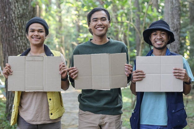 Three young Asian male hikers looking at camera smiling and holding blank cardboard box for text or