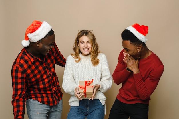 Photo three young and beautiful friends of different nationalities in santa hats are happy with a surprise and a gift box.