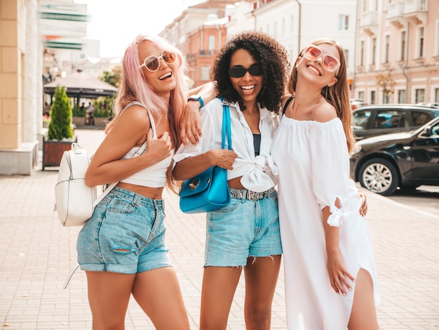 トレンディな夏の服を着た3人の若い美しい笑顔の女性通りの背景でポーズをとるセクシーな屈託のない多民族の女性サングラスで楽しんでいるポジティブモデル陽気で幸せ