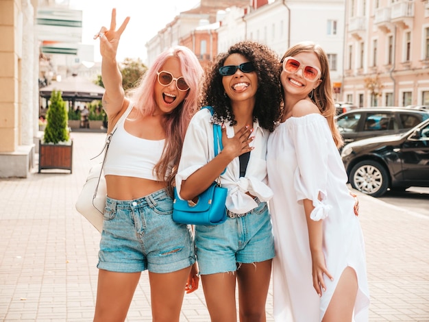 Tre giovani belle donne hipster sorridenti in abiti estivi alla modadonne multirazziali spensierate sexy in posa sullo sfondo della stradamodelli positivi che si divertono con gli occhiali da sole allegri e felici